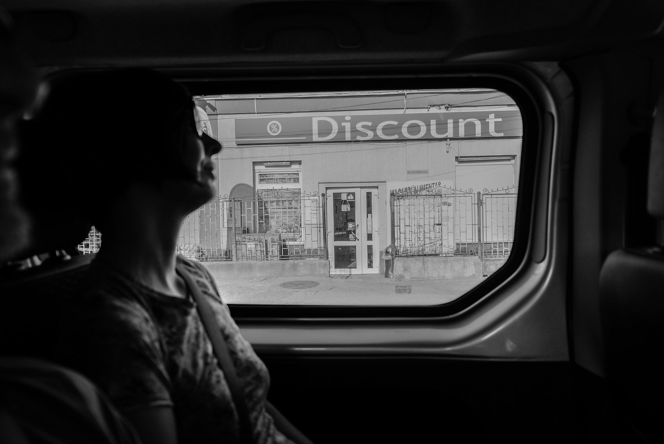 photo en noir et blanc d'une femme dans une voiture regardant par la fenêtre un magasin discount