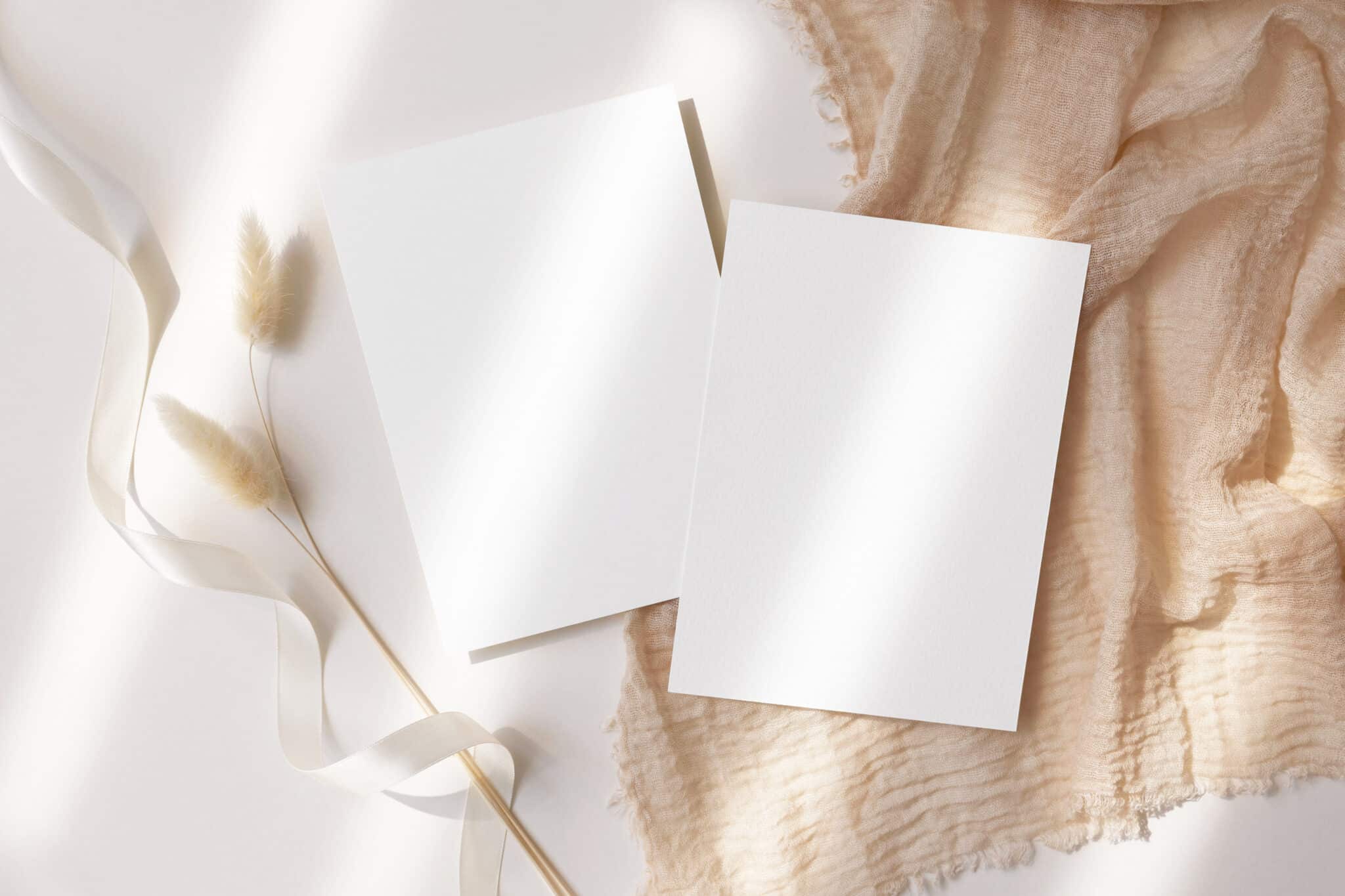 deux feuilles blanches vierges posées sur une table blanche avec un morceau de tissu en lin et une herbe de la pampa et un ruban en soie blanc à côté